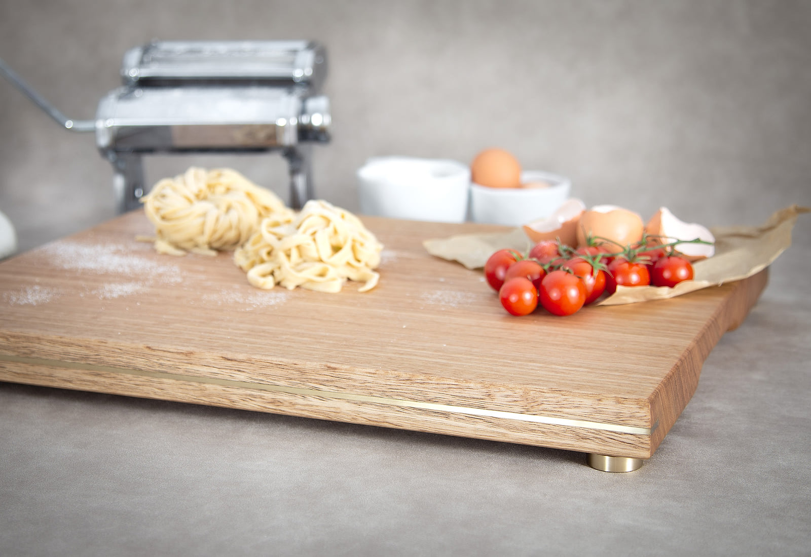 A HEWN chopping and serving board in Tasmanian Oak, brass inlaid along its edge and raised on brass feet, with fresh pasta, cherry tomatoes and eggs